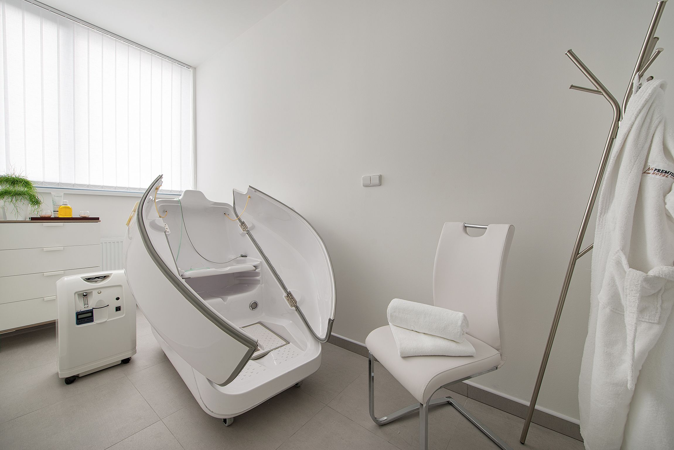 Un cabinet médical moderne, équipé d’un fauteuil inclinable, d’une armoire blanche et d’une fenêtre avec des stores.
