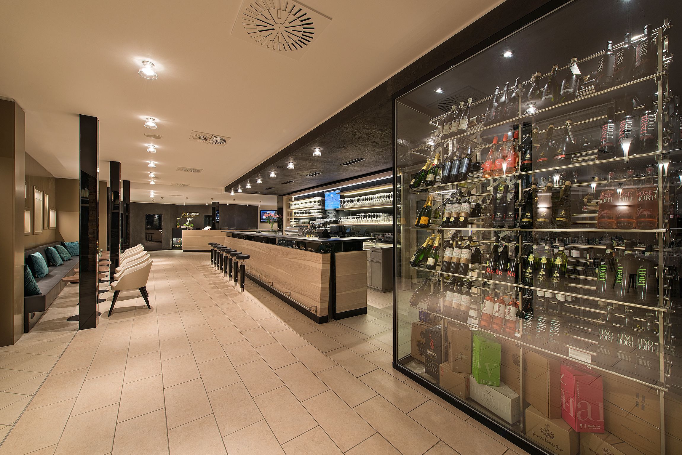Eine moderne Bar mit einer Glastheke voller verschiedener Flaschen und Gläsern, einer Sitzecke mit einer Couch und Sesseln sowie einer Theke mit einem digitalen Bildschirm.