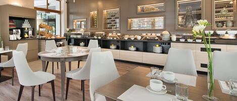 A modern dining area with white chairs and tables, a buffet counter with various food items, and a view of the outdoors through large windows.