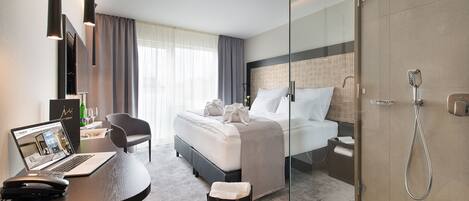 A modern hotel room with a bed, a desk with a laptop, a shower, and a glass partition.