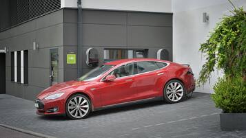 Um Tesla vermelho estacionado em frente a um edifício moderno com fachada cinzenta e sinalização indicando vaga reservada para carros elétricos.