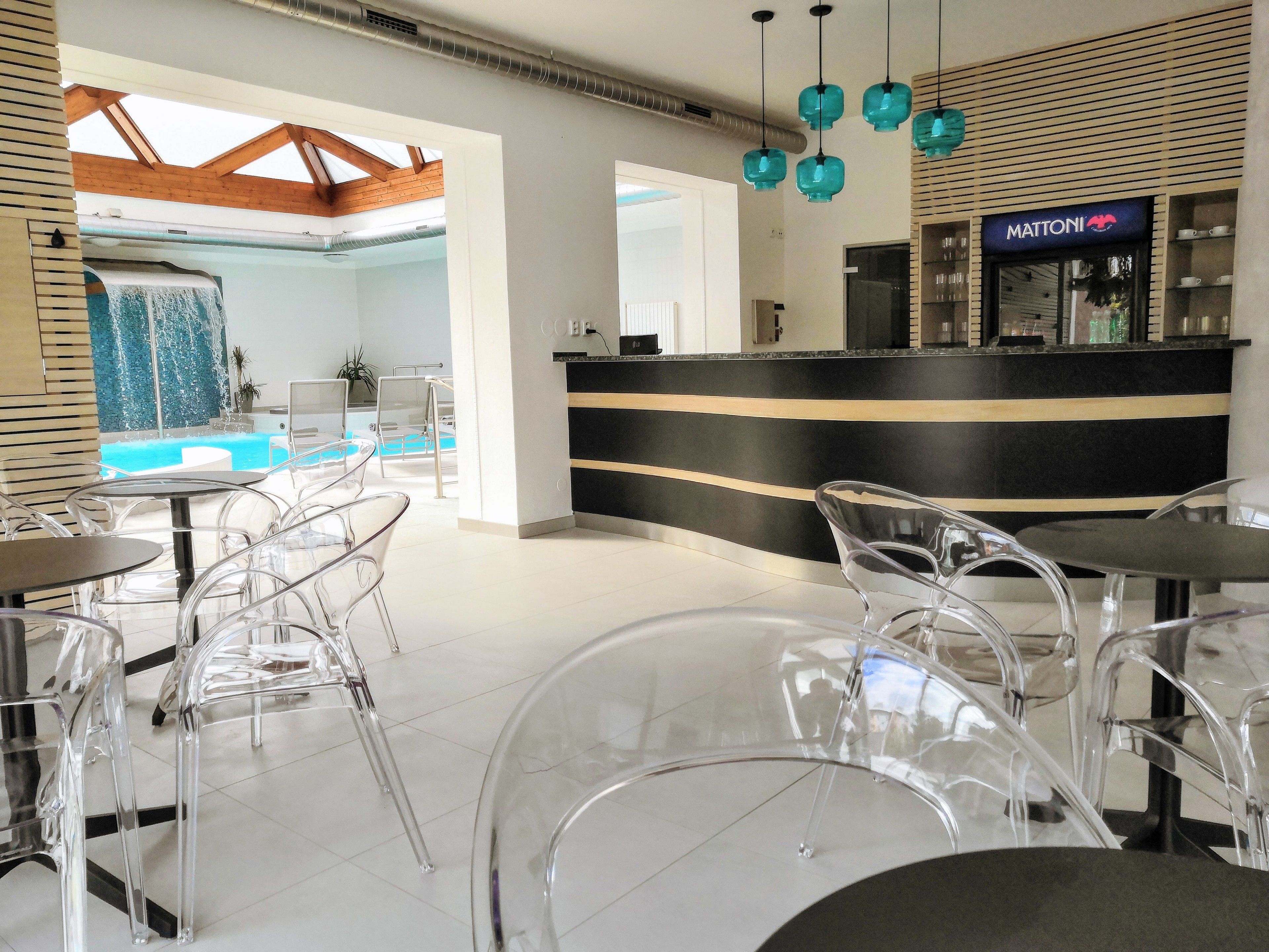 Un moderno vestíbulo de hotel con mostrador de recepción, sillas de plástico transparente, zona de piscina y techo con vigas de madera a la vista.