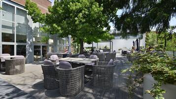Un espace extérieur aménagé avec des meubles en osier et une terrasse en bois, situé à côté d’un bâtiment doté de grandes fenêtres et d’une façade en briques.