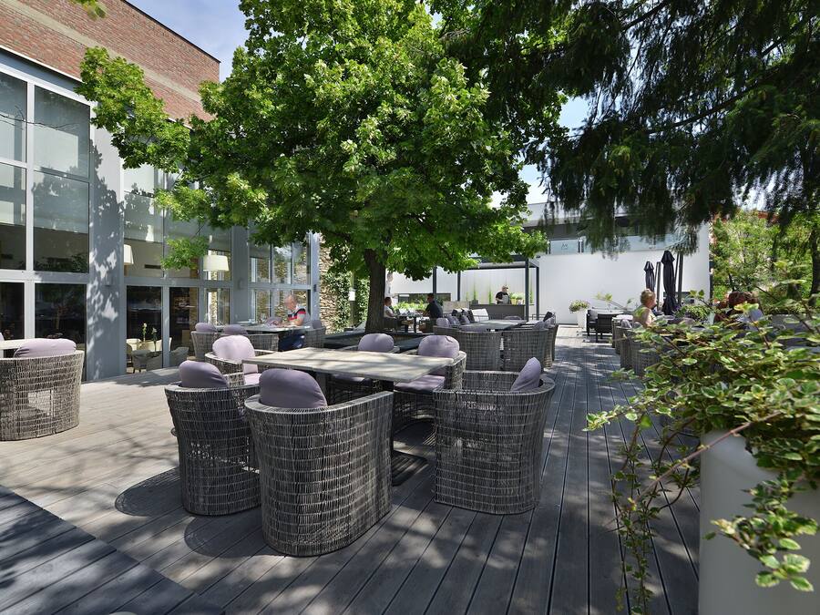 Un espace extérieur aménagé avec des meubles en osier et une terrasse en bois, situé à côté d’un bâtiment doté de grandes fenêtres et d’une façade en briques.