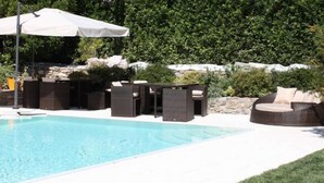 A swimming pool with a white umbrella, wicker furniture, and a stone wall in the background.