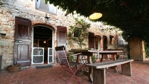 Um edifício rústico de pedra com um banco e mesa de madeira na parte externa, um cesto no chão e um candeeiro pendurado.