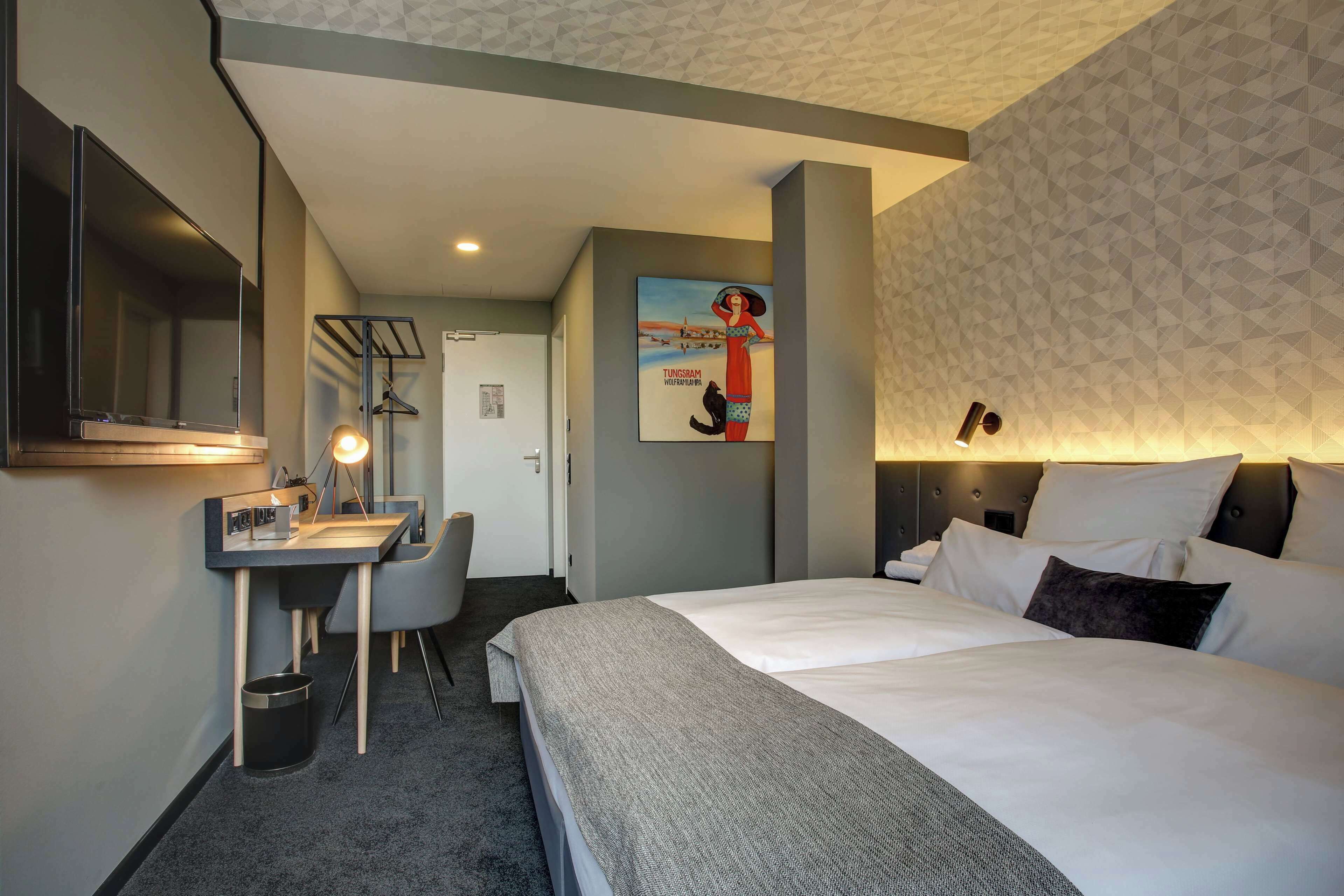 A hotel room with a bed, bedside table, lamp, and a poster on the wall.