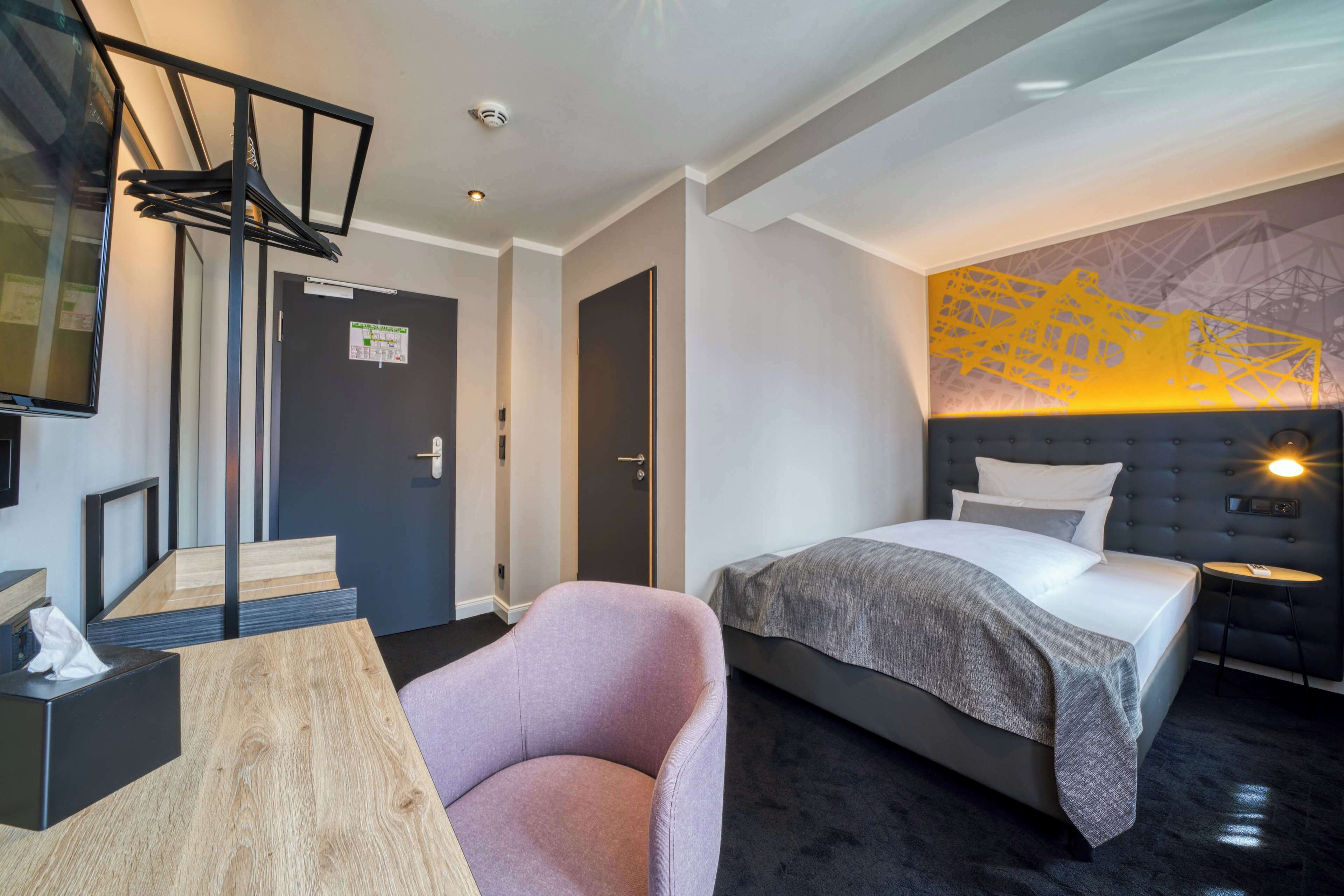Une chambre d’hôtel moderne équipée d’un lit, d’un coin salon avec une table et une chaise, d’un téléviseur fixé au mur et d’une porte avec une pancarte.
