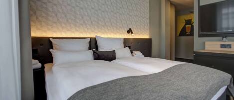 A neatly made bed with white linens and a gray throw blanket, a wall-mounted TV, and a geometric patterned wall.