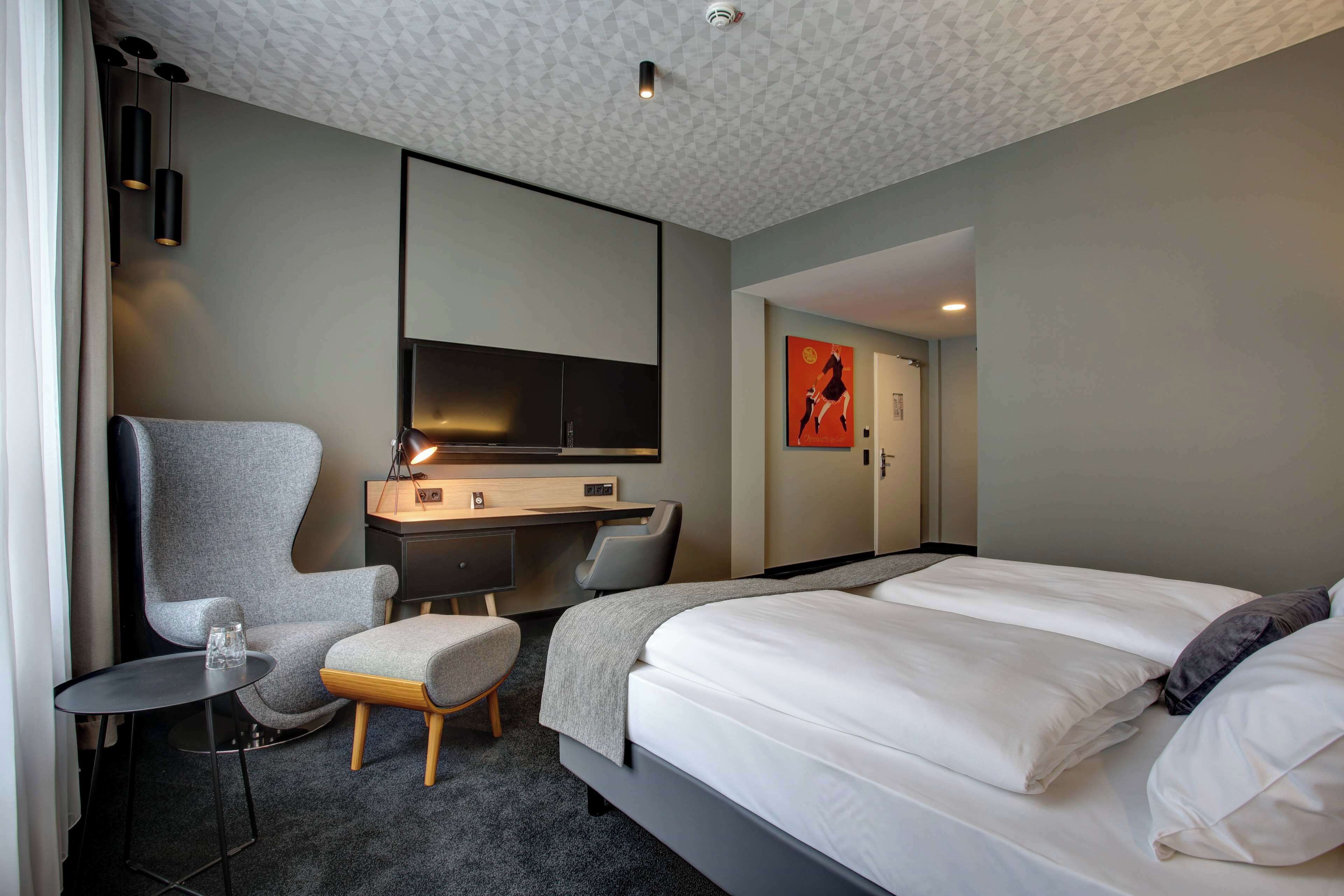 Una habitación de hotel moderna con una cama grande, un escritorio con televisión de pantalla plana, un sillón gris y una mesita.