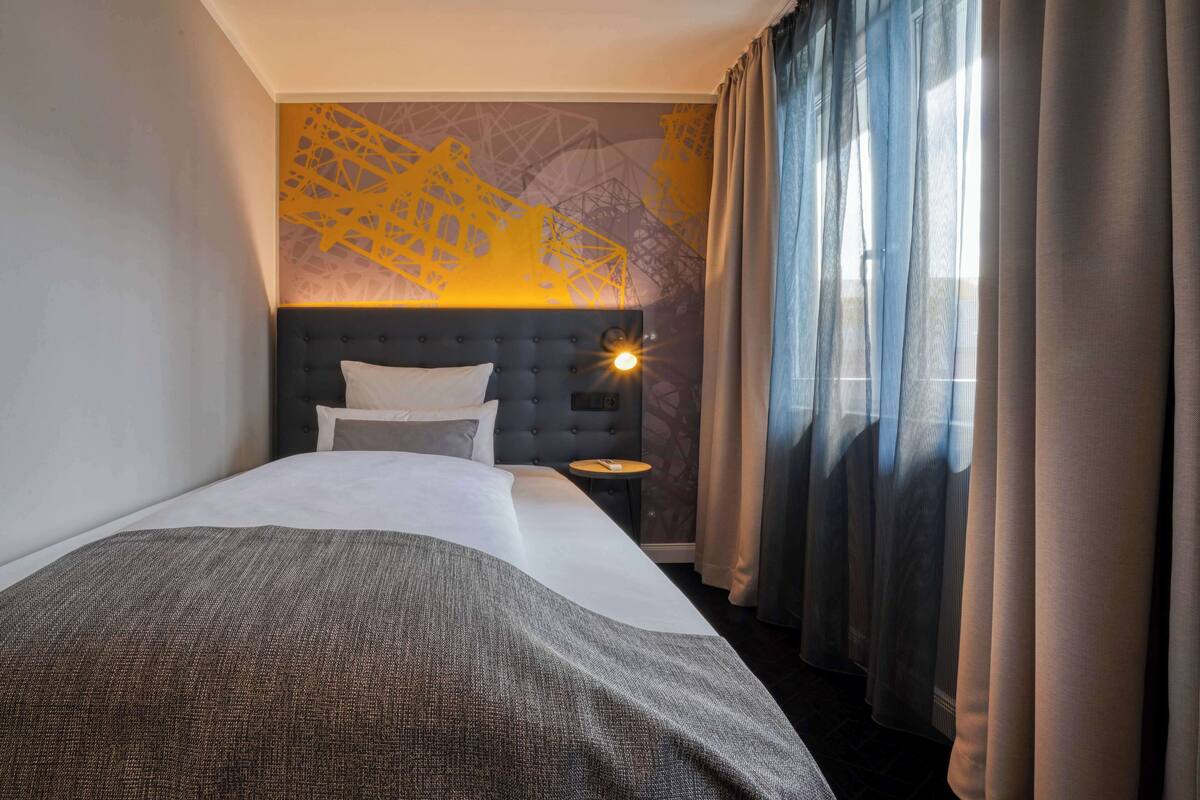 Une chambre d’hôtel avec un grand lit, une tête de lit dotée d’un luminaire, une petite table et des rideaux légers.