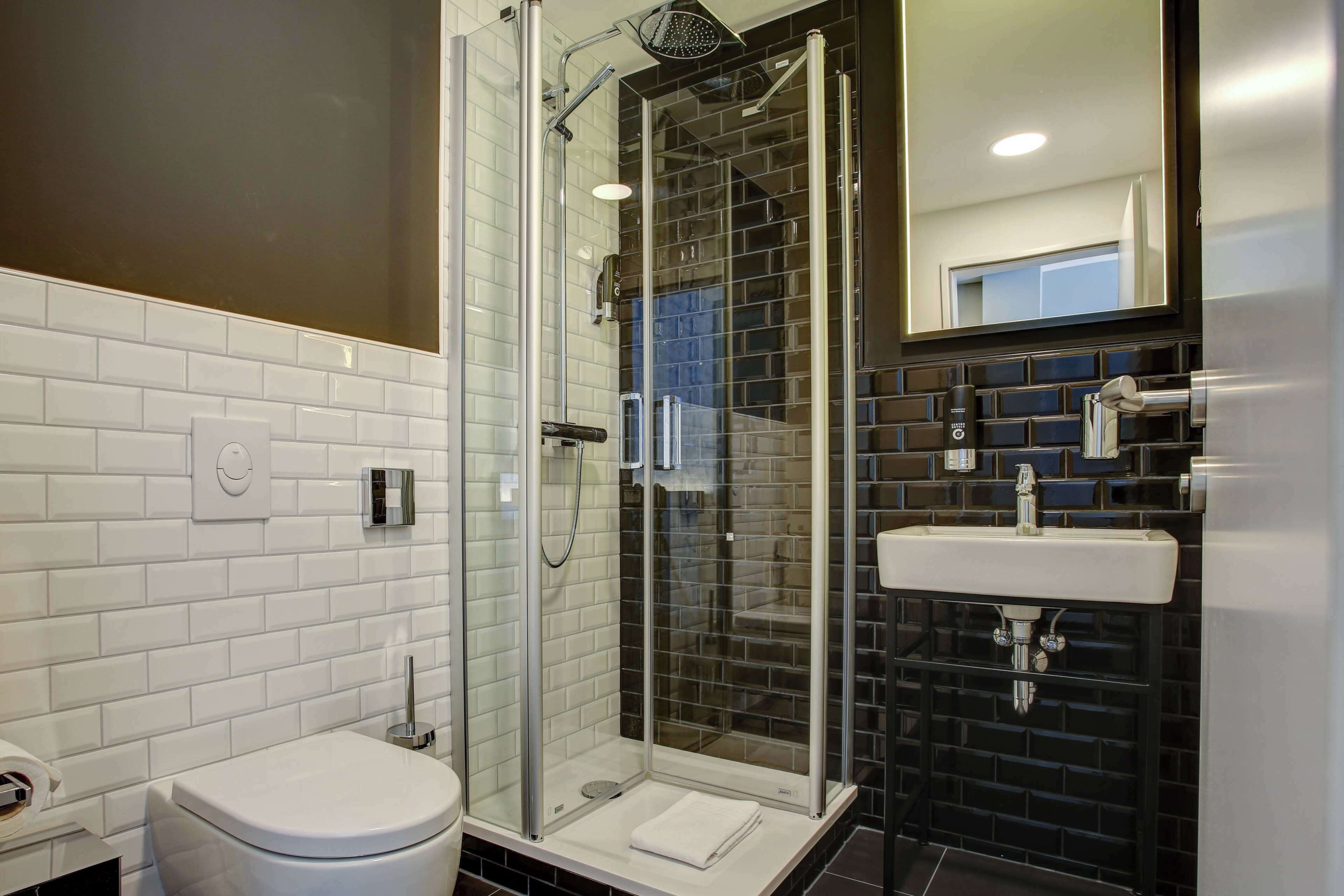 Un baño moderno con ducha envuelta en vidrio, inodoro blanco y lavamanos con zócalo de azulejos negros.