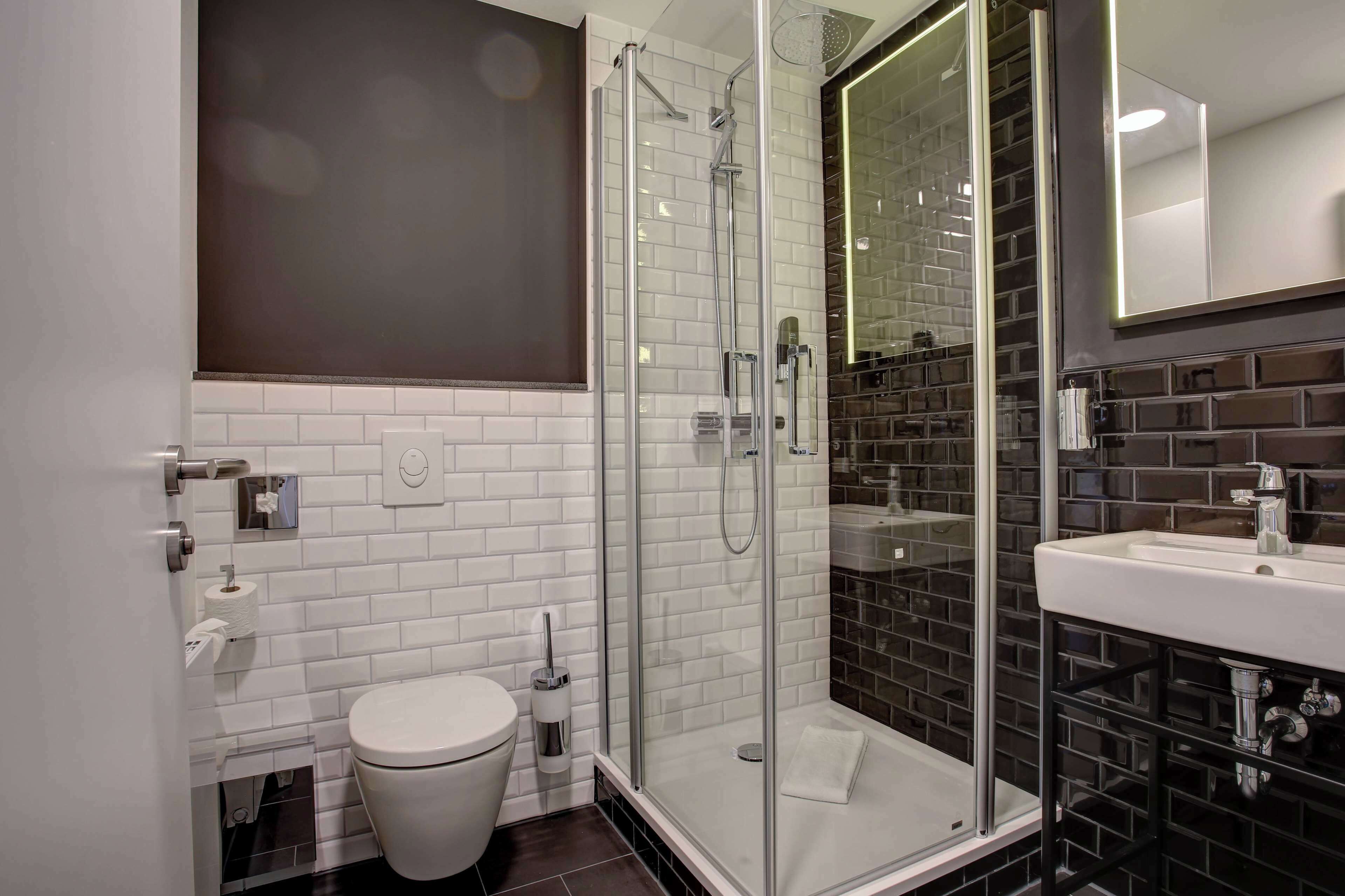 Une salle de bain moderne avec une douche à parois de verre, des murs recouverts de carreaux blancs, des toilettes blanches et un lavabo dont le plan est recouvert de carreaux noirs.