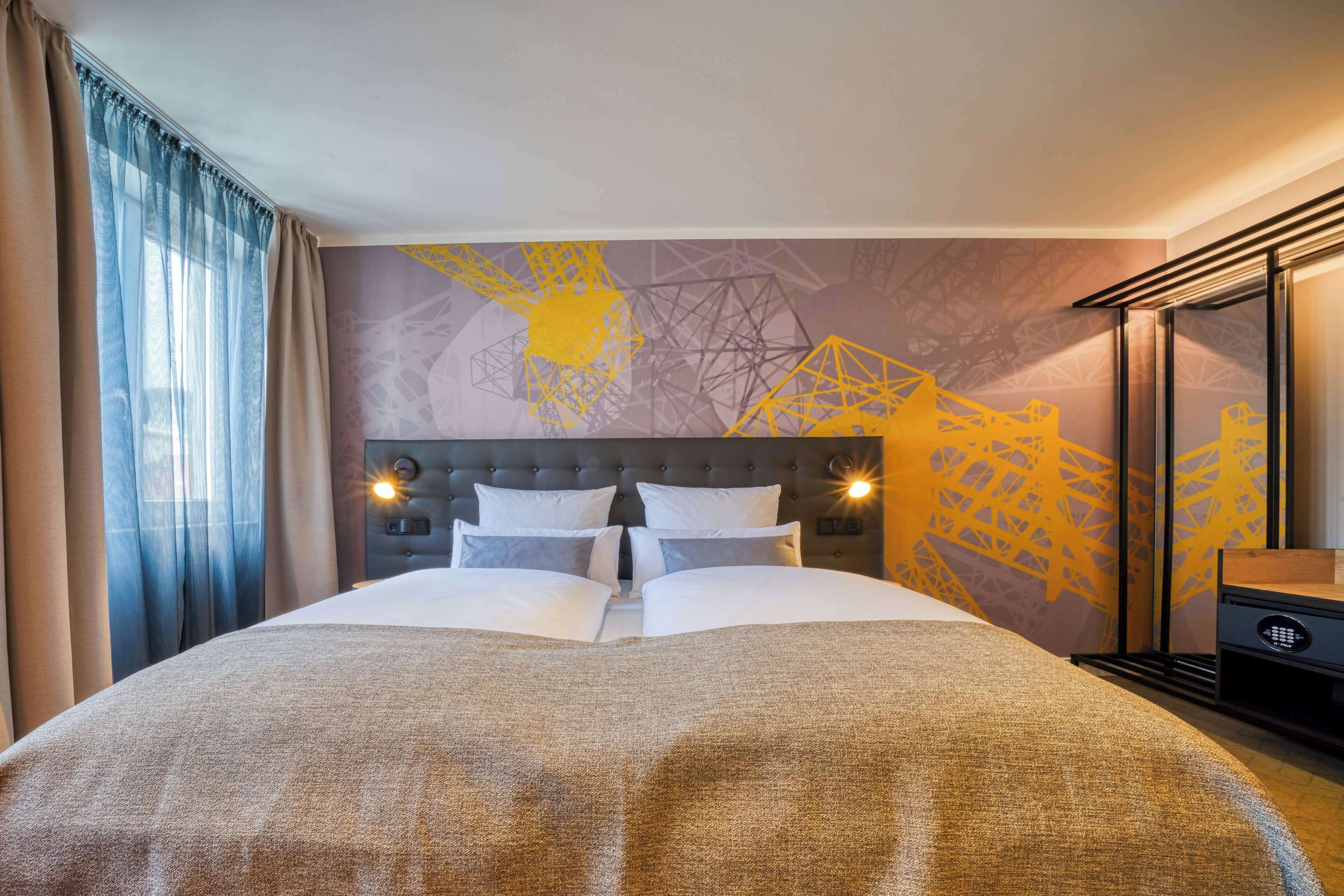 Quarto de hotel com cama grande, cabeceira com iluminação embutida, mesa de cabeceira e parede com padrão geométrico.