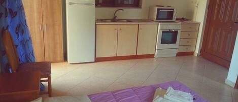 A bedroom with a bed, a purple blanket, a wooden wardrobe, a white refrigerator, a microwave, and a small oven.