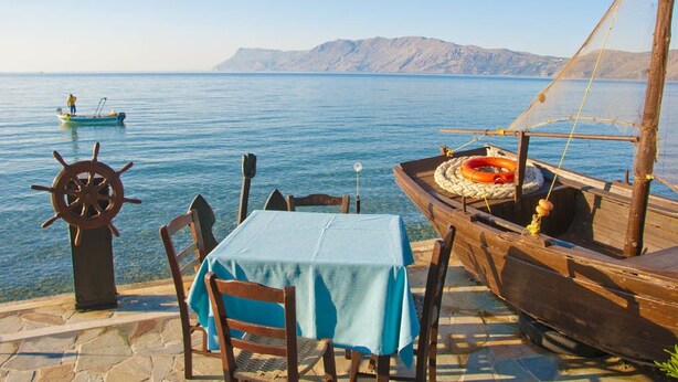 A wooden boat with a table and chairs by the water, a steering wheel, and a lifebuoy. - Akrogiali (Kissamos)
