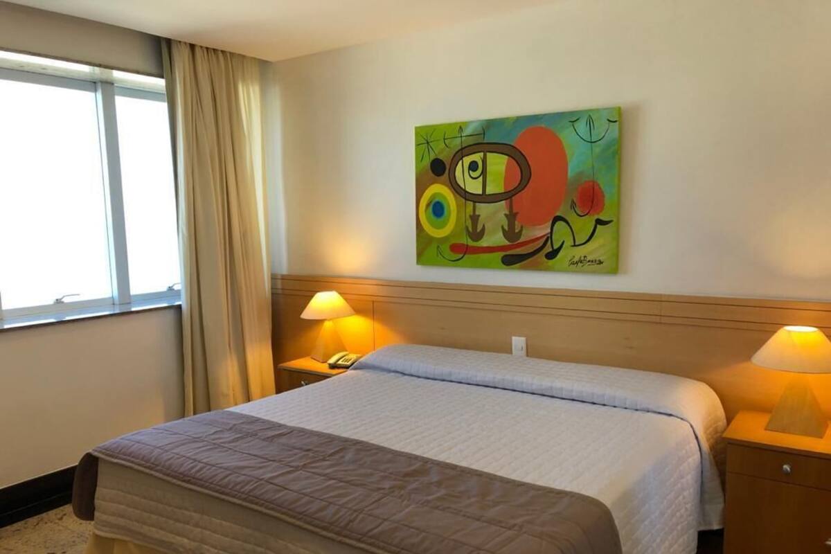 A hotel room with a bed, two bedside lamps, a painting on the wall, and a window with curtains.