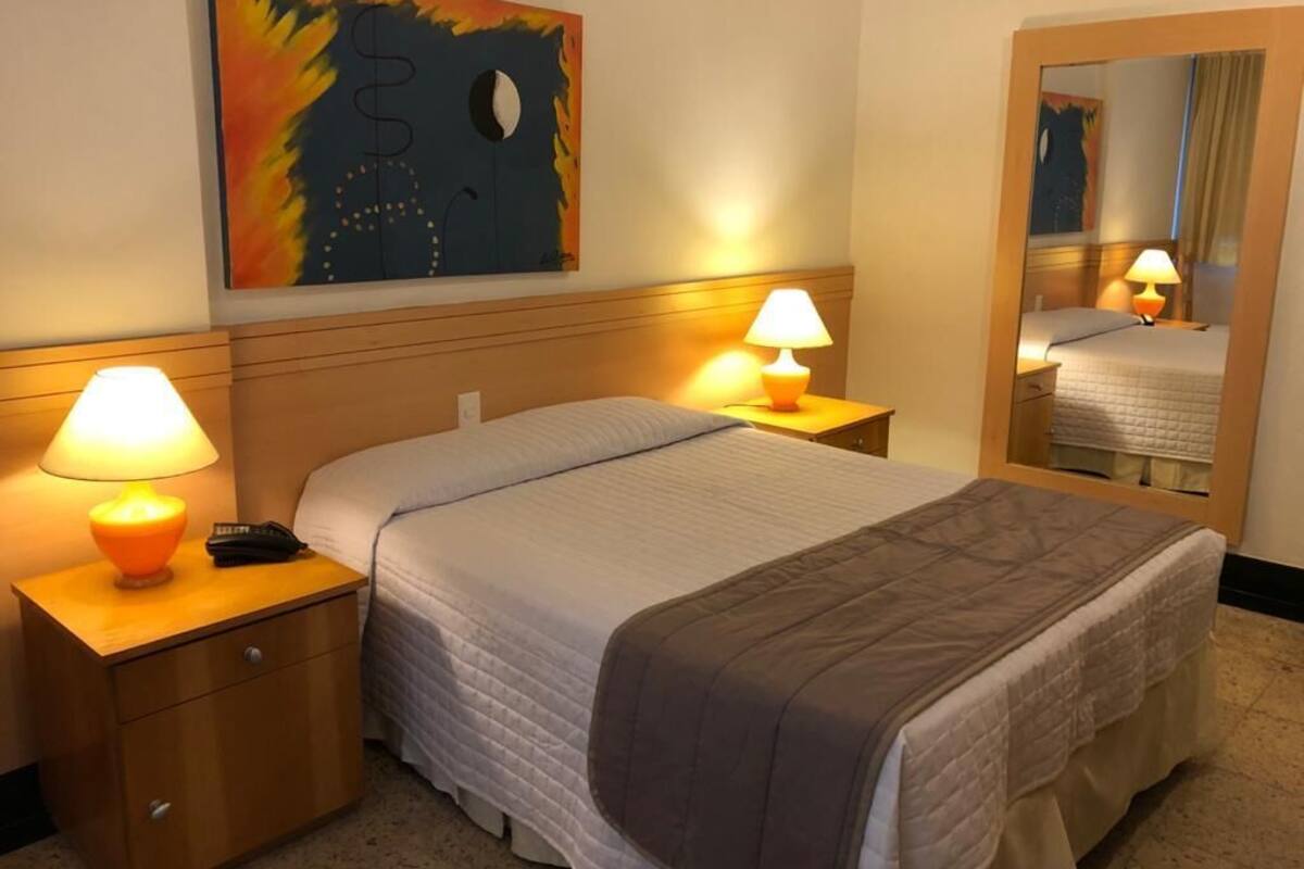 A hotel room with a bed, bedside lamps, a nightstand, a mirror, and a painting on the wall.