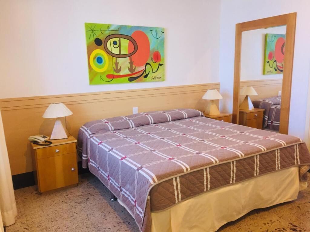 Quarto de hotel com cama, mesas de cabeceira, luminárias, espelho e um quadro abstrato colorido na parede.