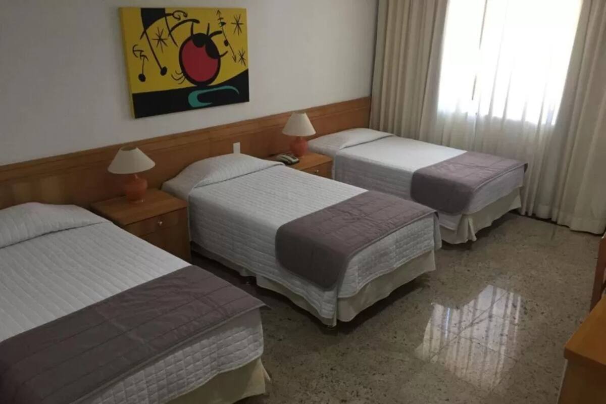A hotel room with three beds, a wooden headboard, a painting on the wall, and a window with curtains.