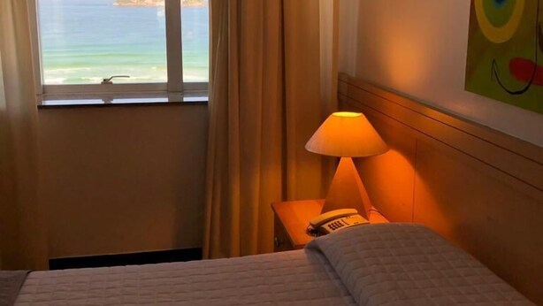 A hotel room with a bed, a nightstand with a lamp, a window with a view of the sea, and a painting on the wall. - Tropical Barra Hotel (Rio de Janeiro)