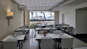 A restaurant with a view of the sea, featuring a modern interior with white tables and chairs, a patterned ceiling, and a large window. - Tropical Barra Hotel (Rio de Janeiro)