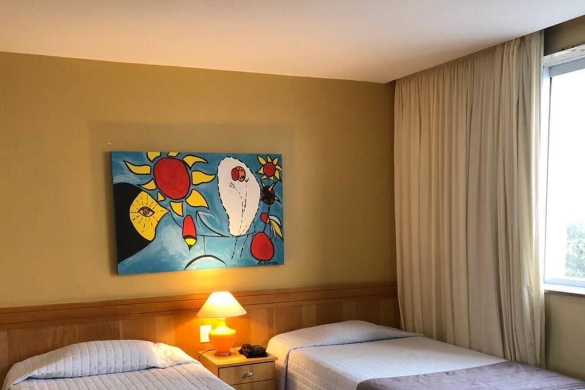 A hotel room with two beds, a nightstand with a lamp, a painting on the wall, and a window with curtains.