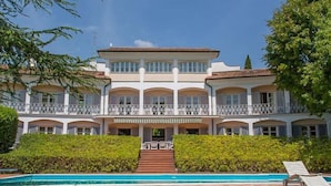 Un edificio ampio e a più piani con una scala centrale che conduce a un balcone, circondato da vegetazione lussureggiante e una piscina.