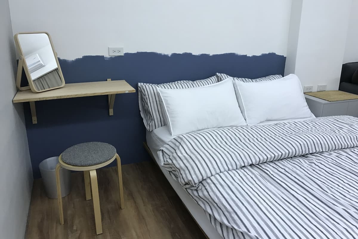 A bedroom with a bed, a small stool, a shelf, and a wall socket.