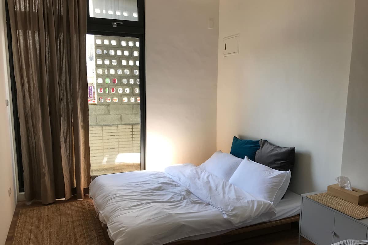 A bedroom with a bed, a nightstand, a window with curtains, and a view of the outside.
