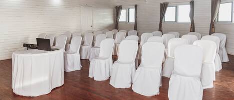 A room with wooden floors, white walls, and a wooden ceiling. There are rows of white chairs with skirts, a laptop on a table, and a door.