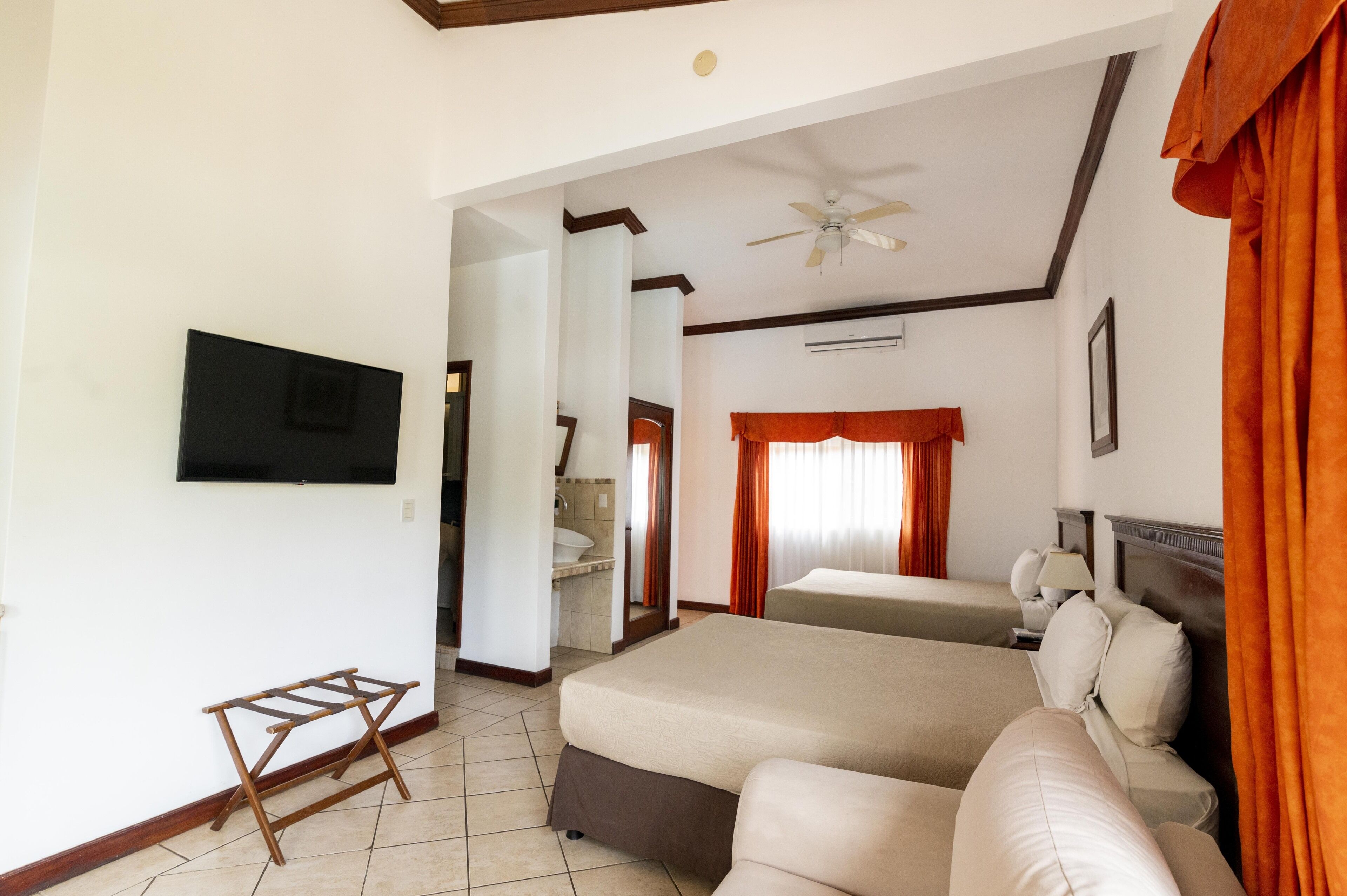 Une chambre d’hôtel avec un grand lit, un lit plus petit, un téléviseur fixé au mur, un ventilateur de plafond et une salle de bain visible à travers une porte ouverte.