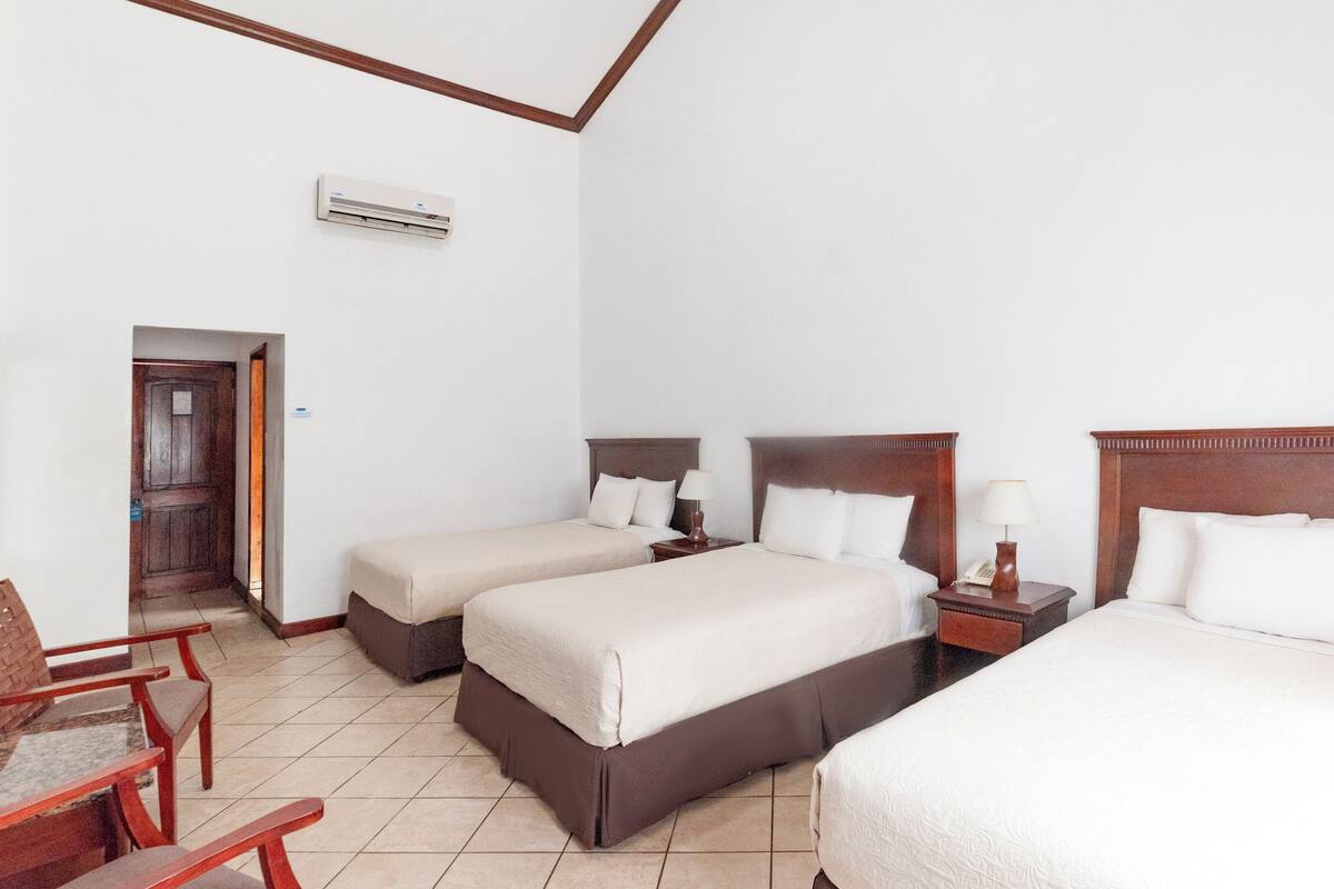 A hotel room with three beds, wooden furniture, and an air conditioning unit.