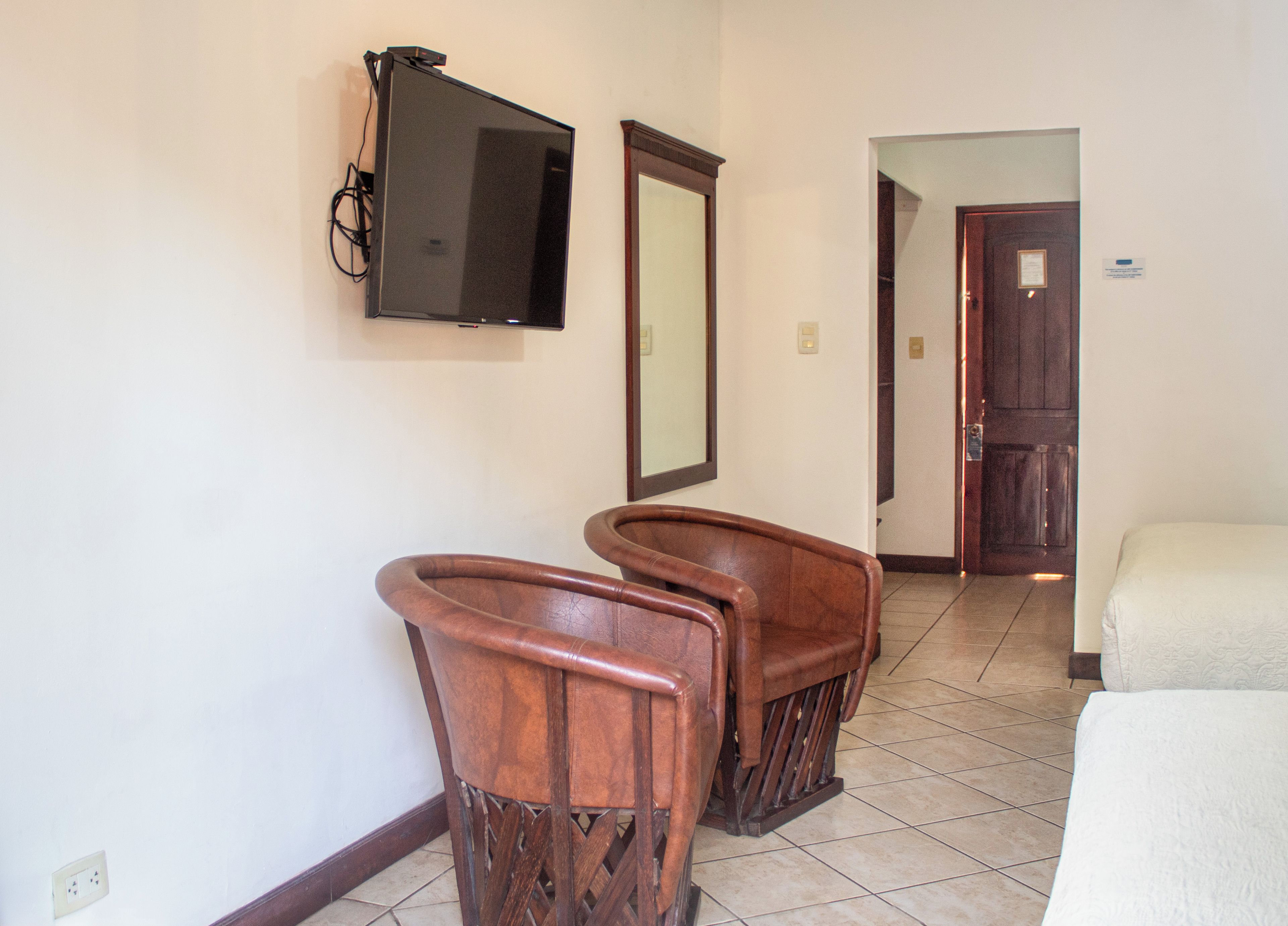 Una habitación de hotel con una televisión montada en la pared, una silla de madera, una cama y una puerta que da a otra habitación.