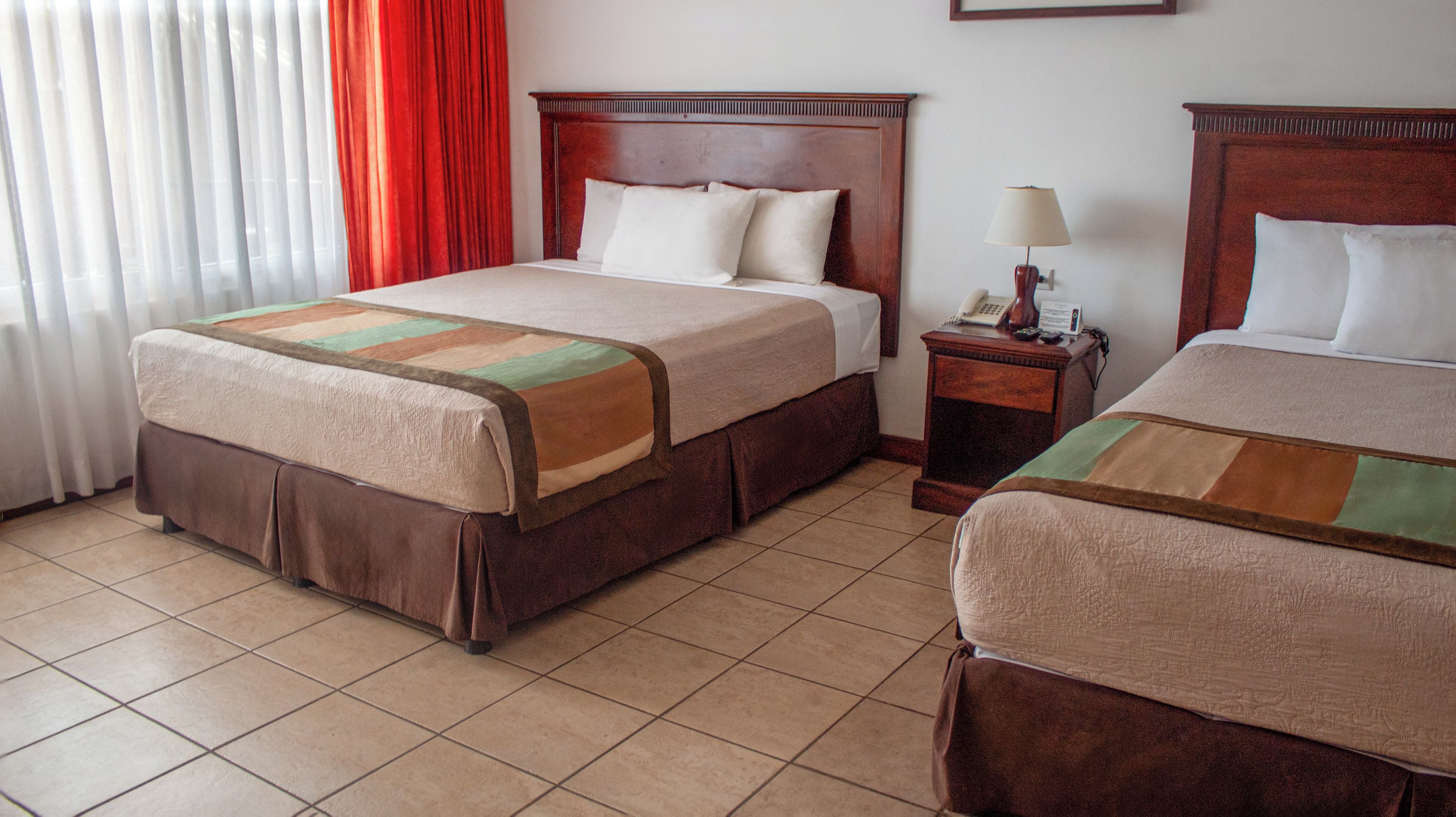 Une chambre d’hôtel avec deux lits, une table de chevet avec une lampe et une fenêtre avec des rideaux.