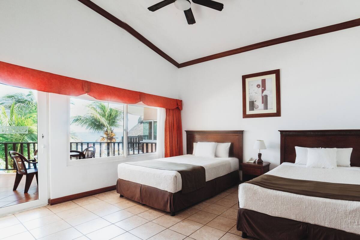 A hotel room with two beds, a balcony with chairs, and a ceiling fan.
