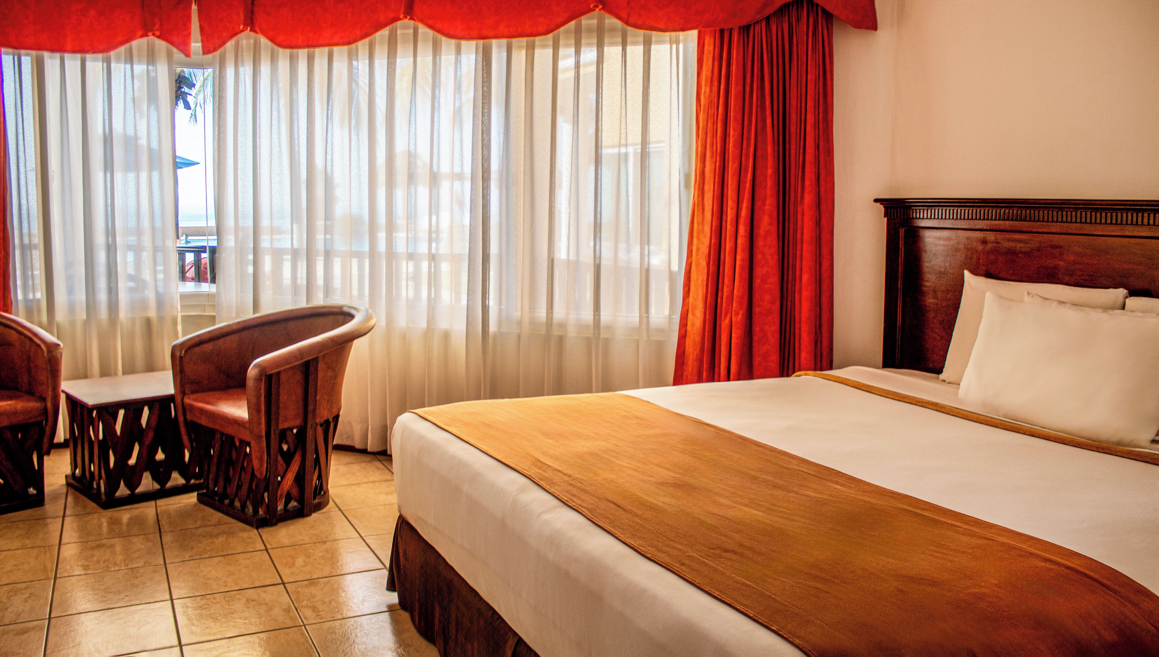 A hotel room with a bed, a chair, a small table, and a view through sheer curtains.
