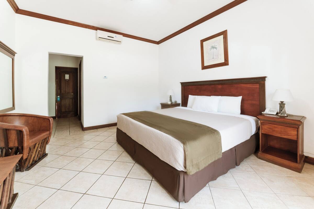 A hotel room with a large bed, a wooden headboard, a nightstand with a lamp, a desk with a chair, and a door leading to another room.
