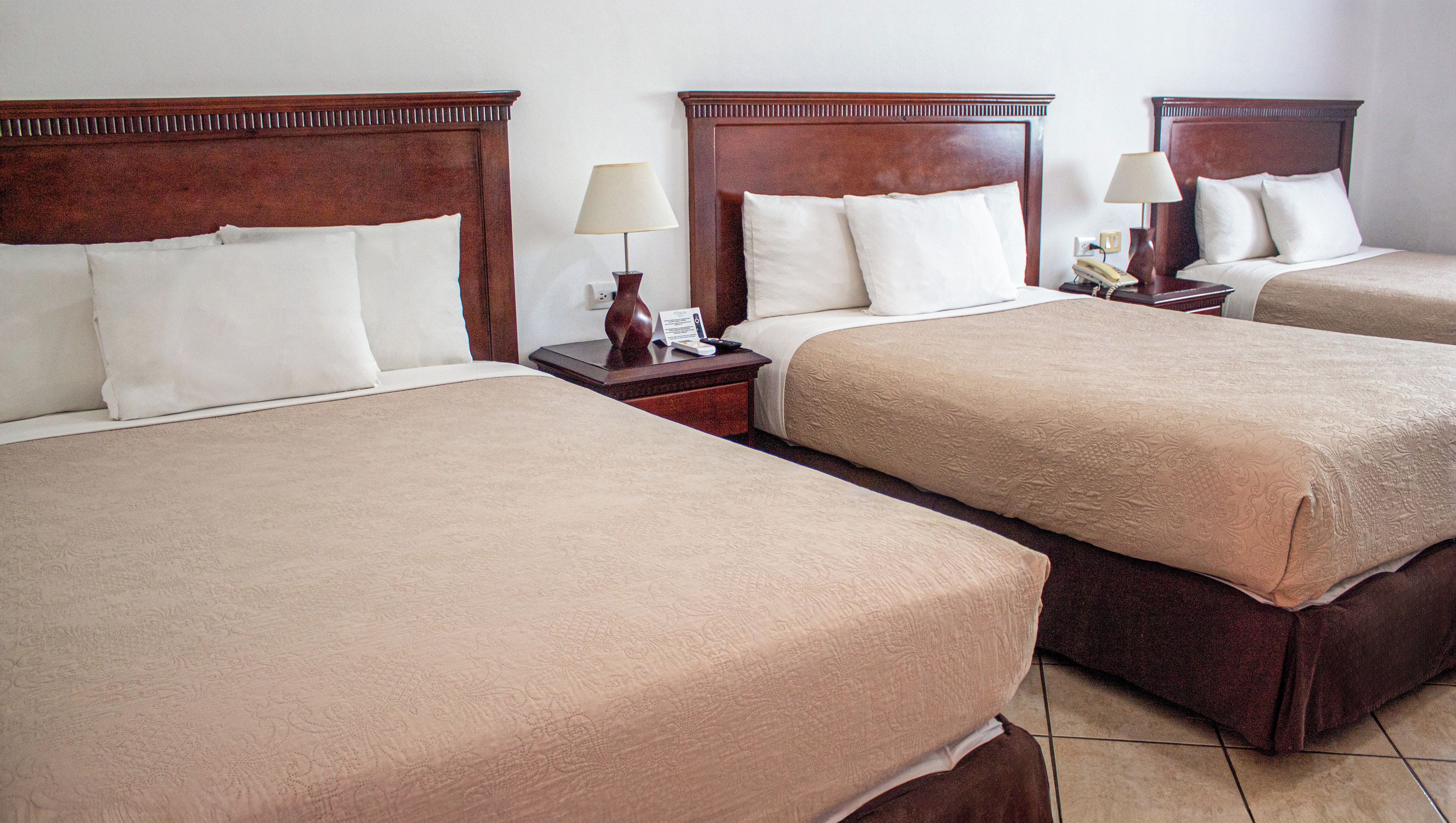 A hotel room with three double beds, each with a wooden headboard, a bedside table with a lamp, and a nightstand with a phone.
