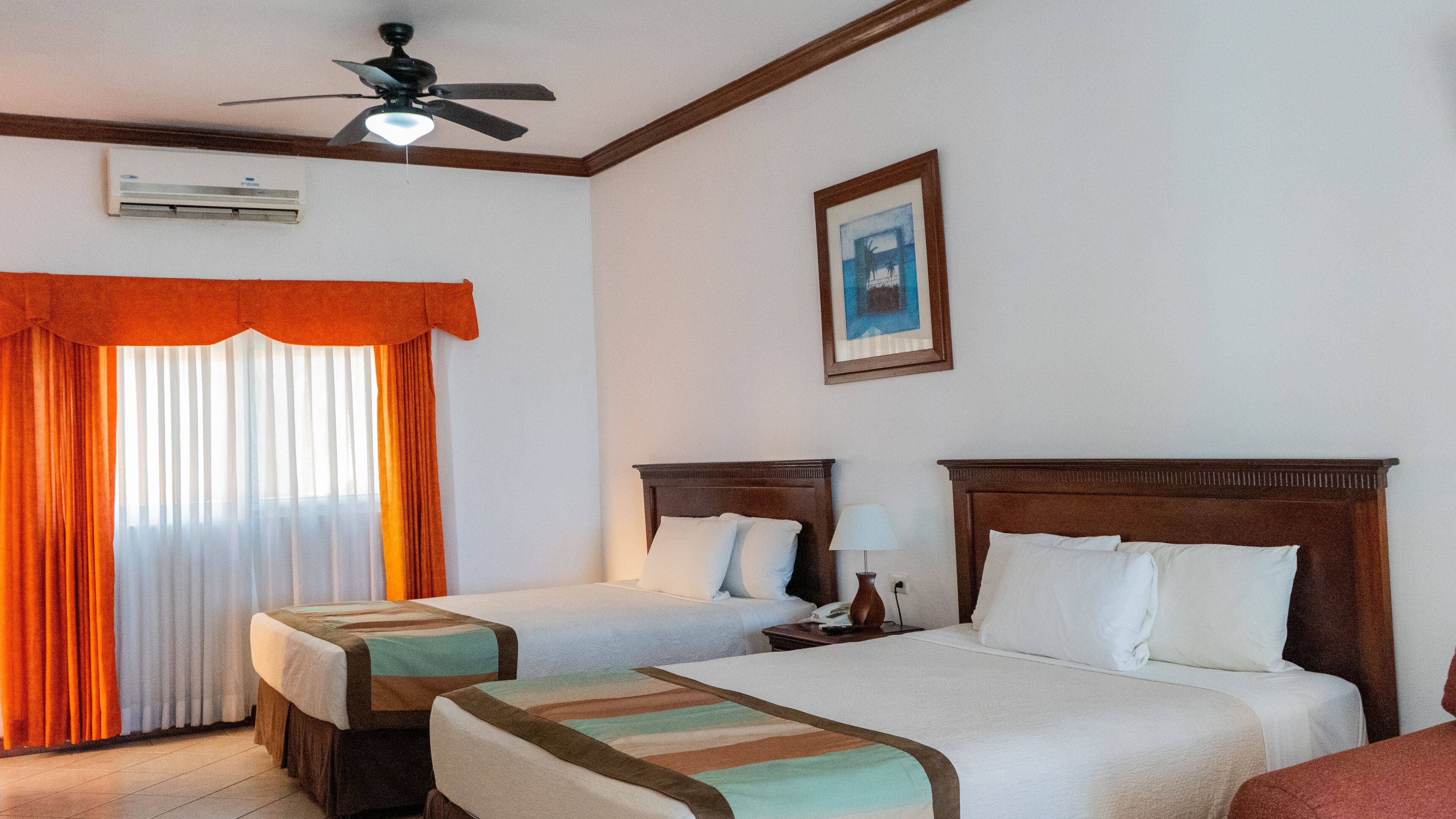Una habitación de hotel con dos camas, un ventilador de techo, un equipo de aire acondicionado y un cuadro en la pared.