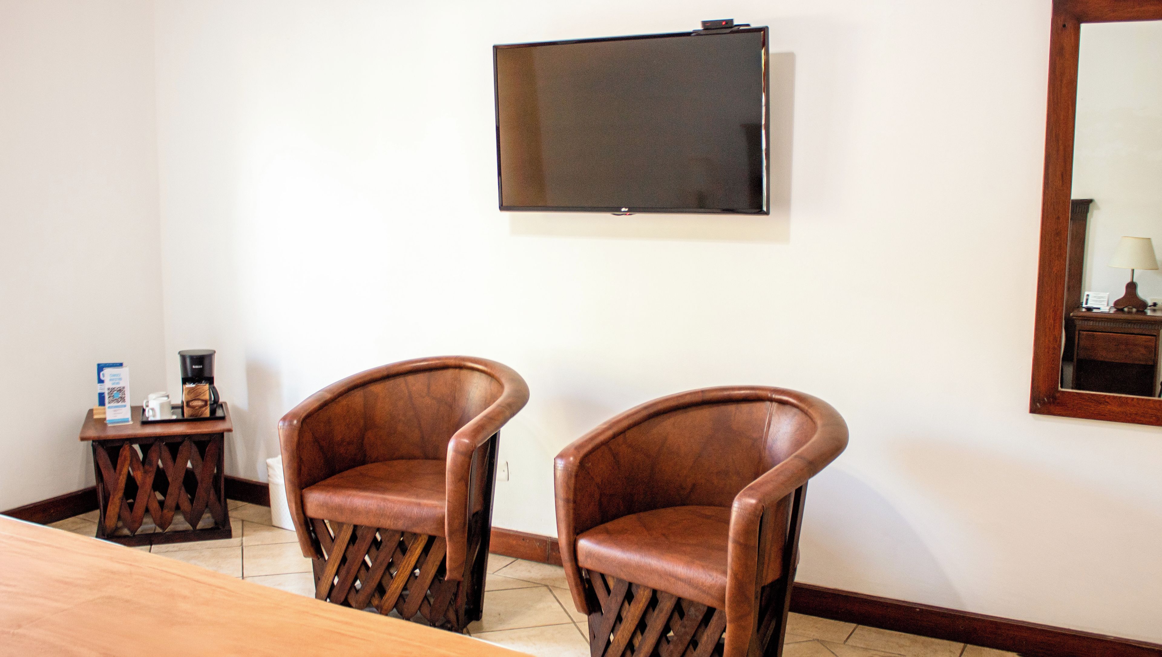 A room with two wooden chairs, a coffee station, a flat-screen TV mounted on the wall, and a wooden table.