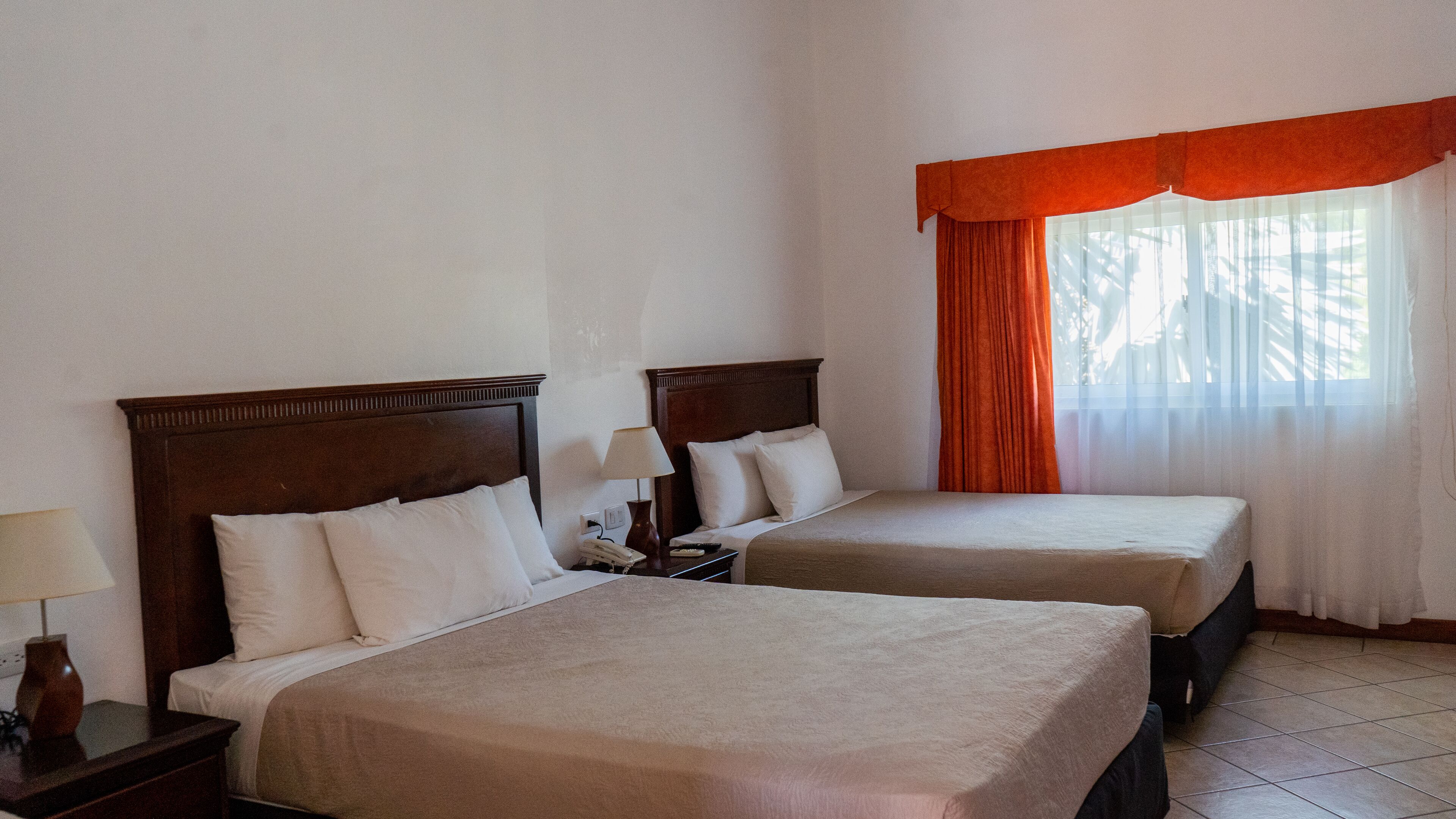 A hotel room with two beds, a nightstand with a lamp, a telephone, and a window with orange curtains.