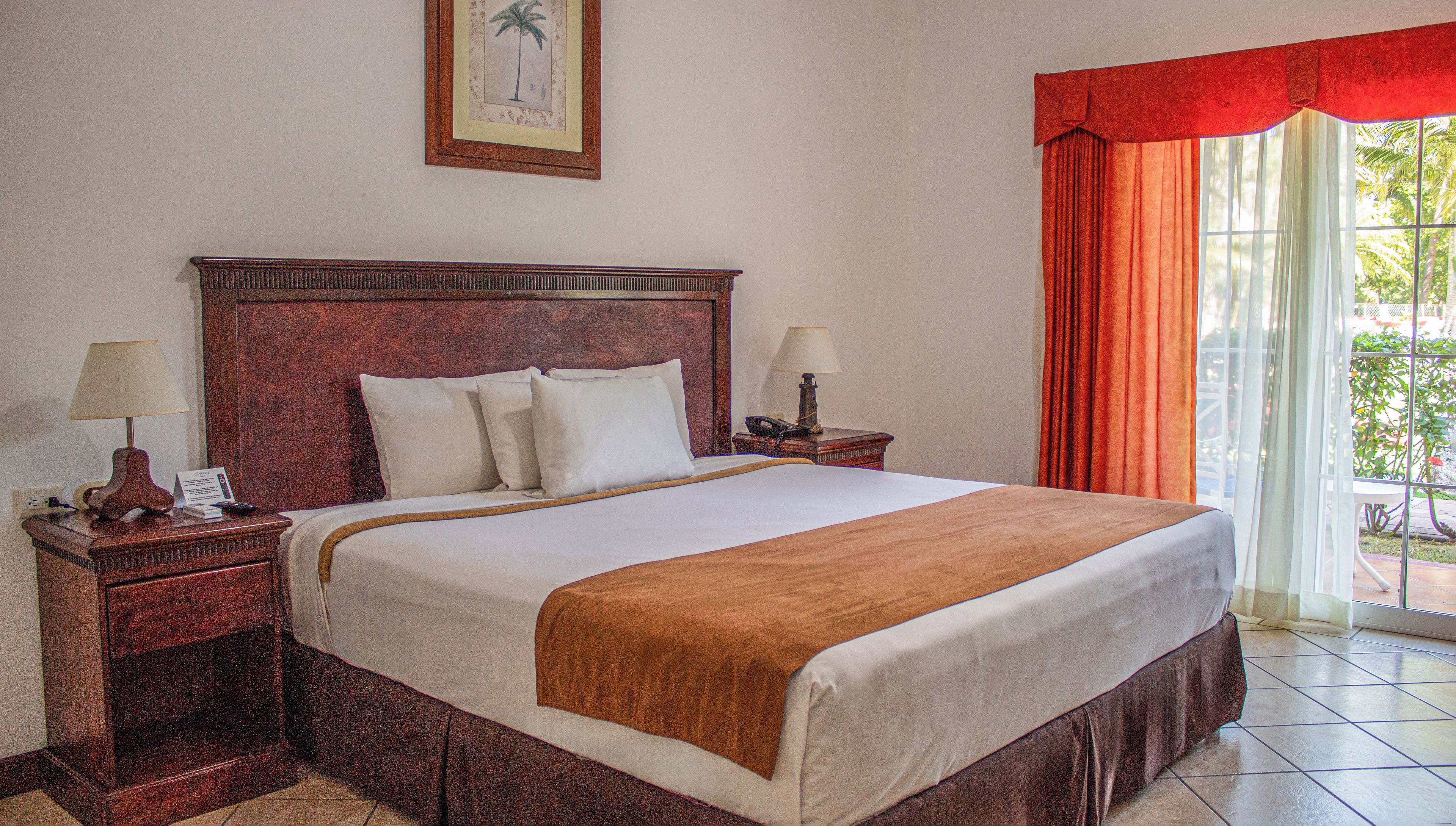 Une chambre d’hôtel avec un grand lit, deux tables de chevet, une petite table, une lampe, un tableau encadré au mur et une fenêtre avec des rideaux.