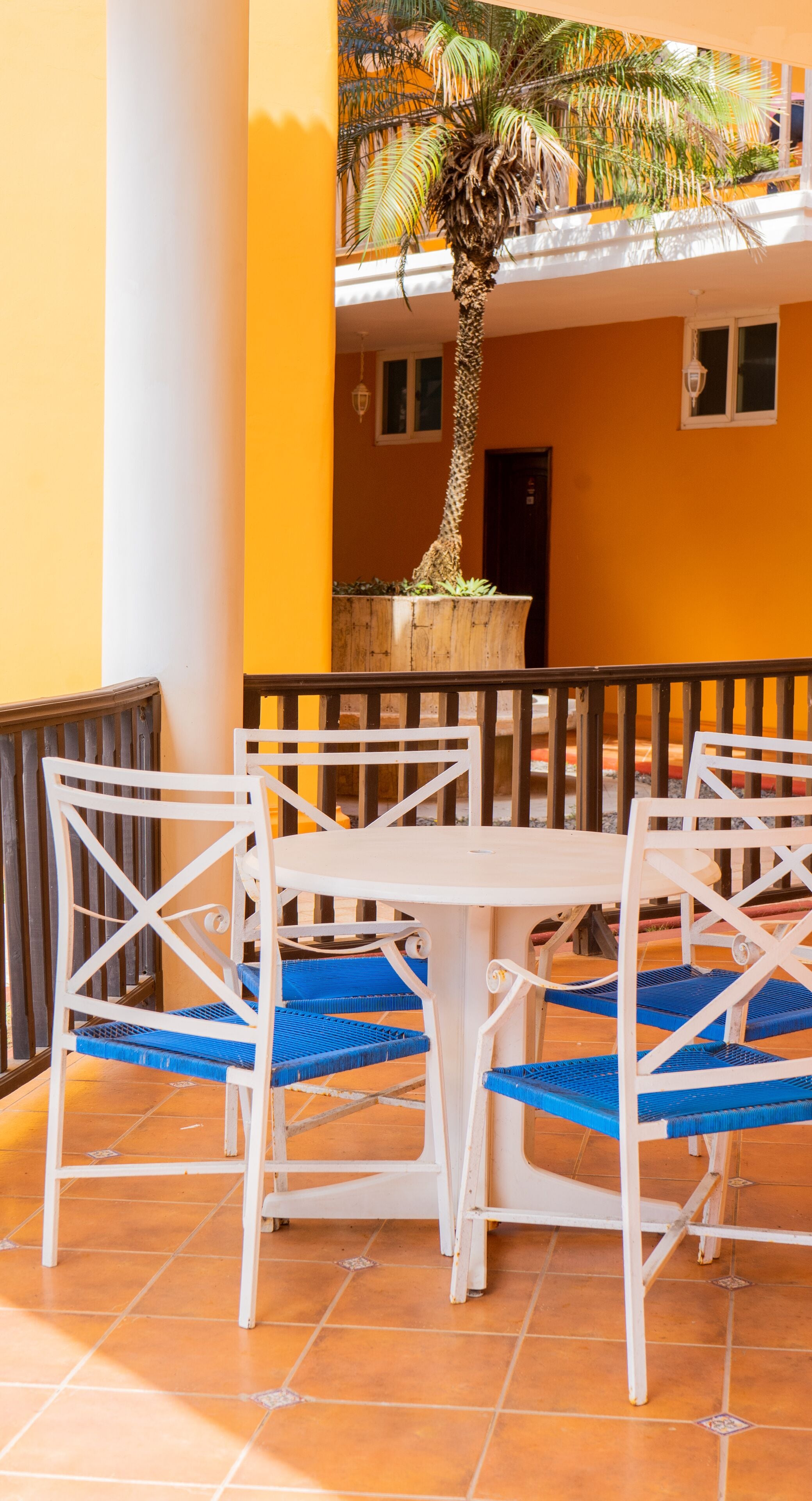 A terrace with a white table and blue chairs, a palm tree, and a yellow building in the background.