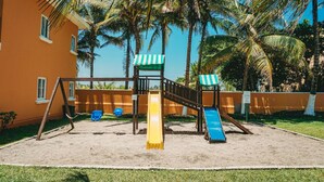Un parque infantil con tobogán y estructura de escalada, rodeado de palmeras y un edificio de fachada amarilla.