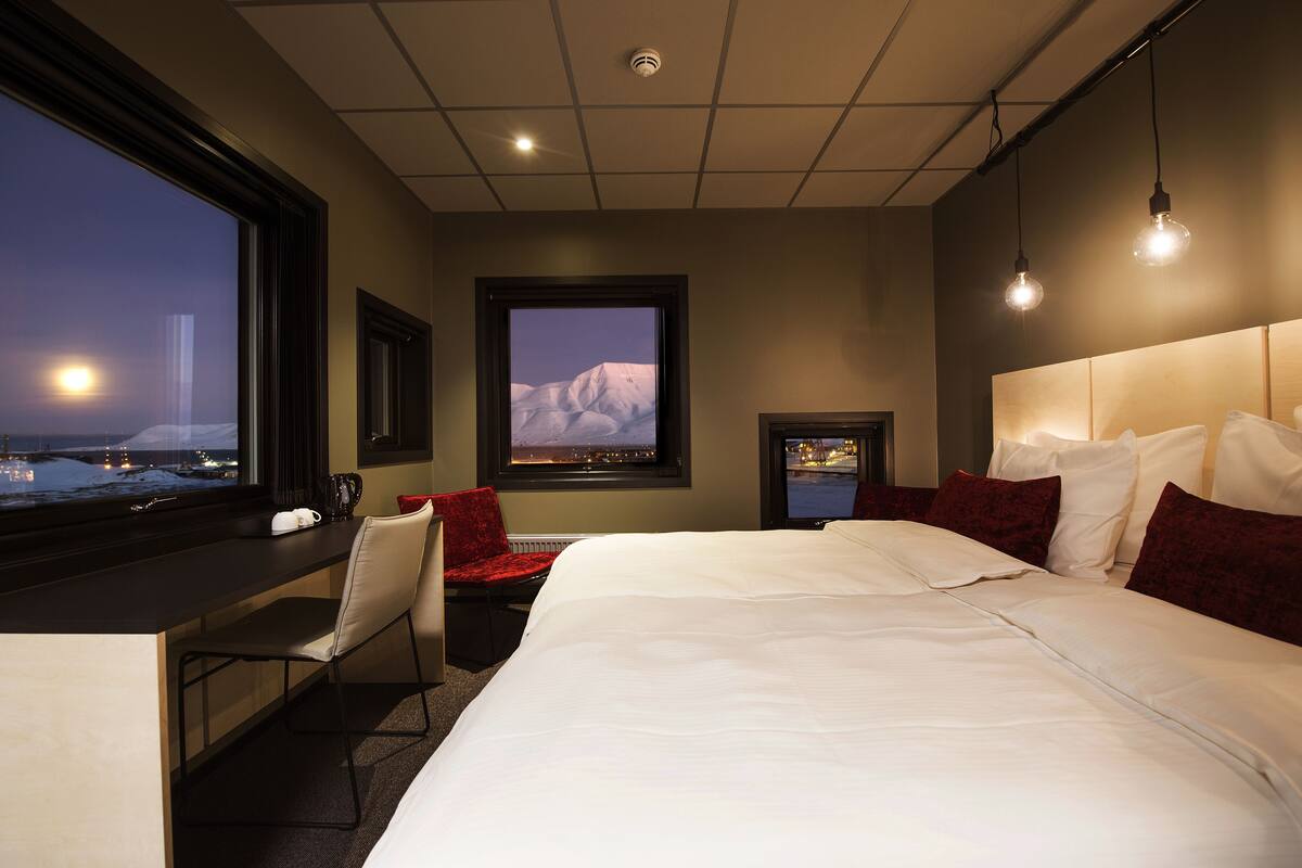 A hotel room with a large bed, a desk, a chair, and a window with a view of a snowy landscape.