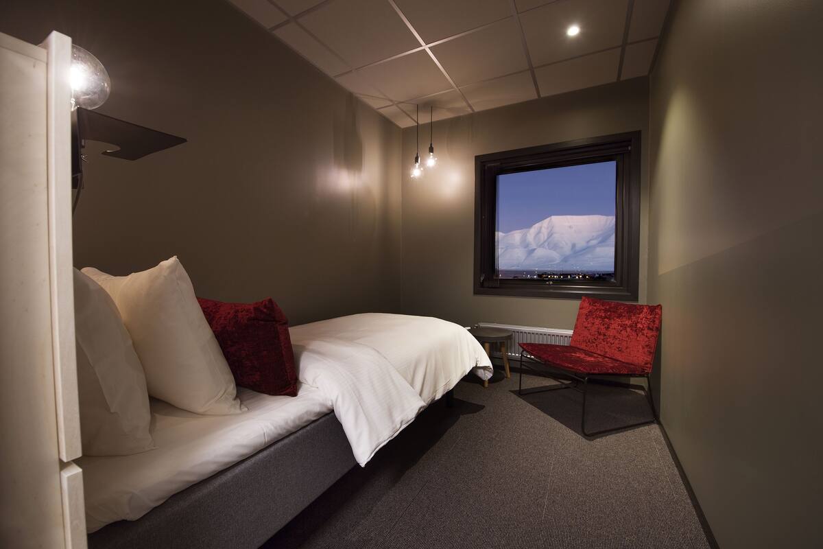 A hotel room with a bed, a red chair, a TV on the wall, and a window with a snowy mountain view.