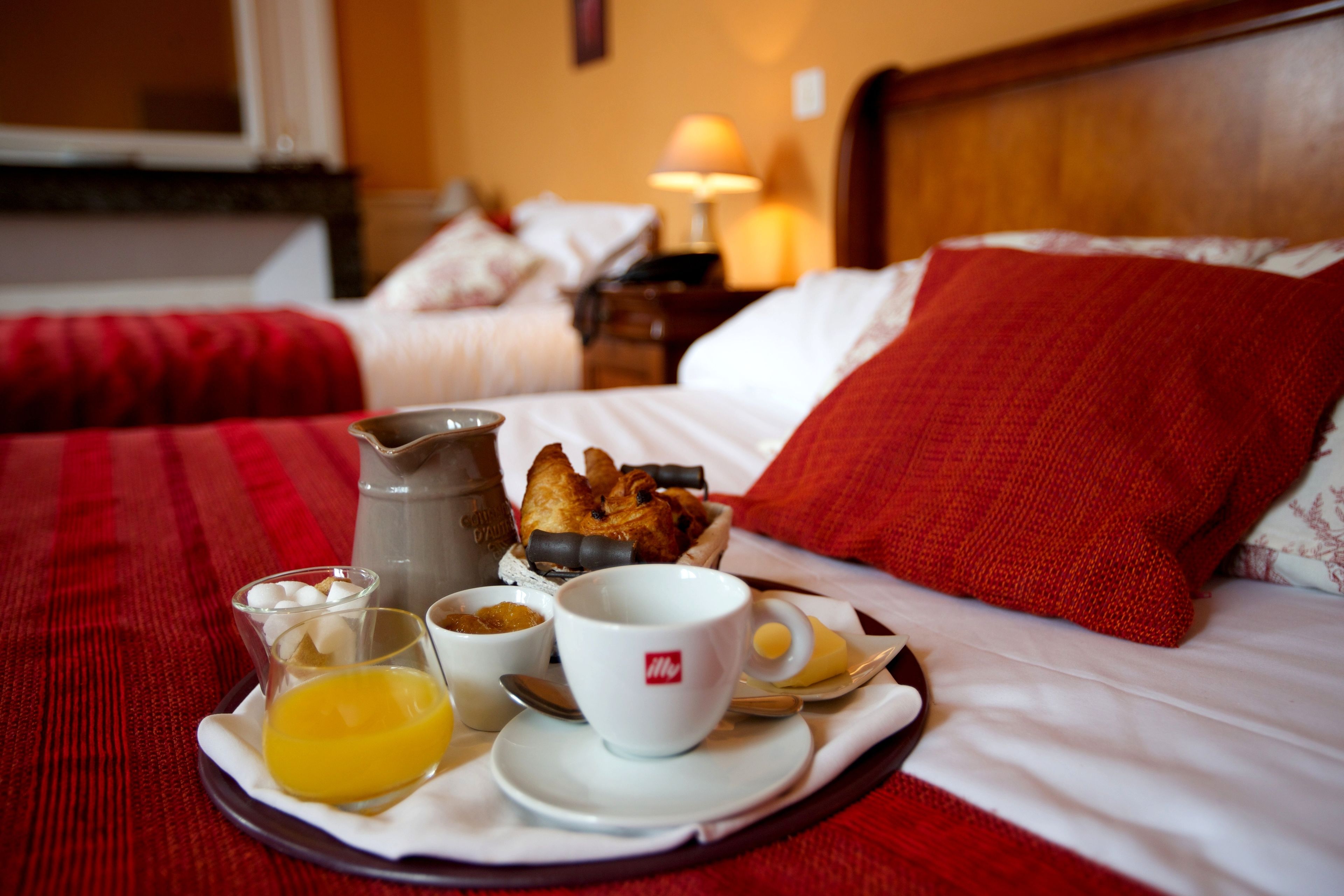 A breakfast tray with coffee, croissants, and orange juice on a bed with red bedding and a wooden headboard.