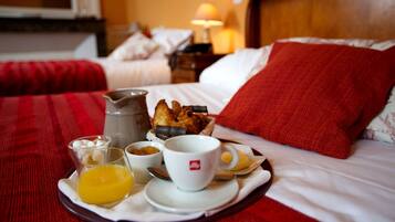 Bandeja de café da manhã com café, croissants e suco de laranja sobre uma cama com roupa de cama vermelha e cabeceira de madeira.