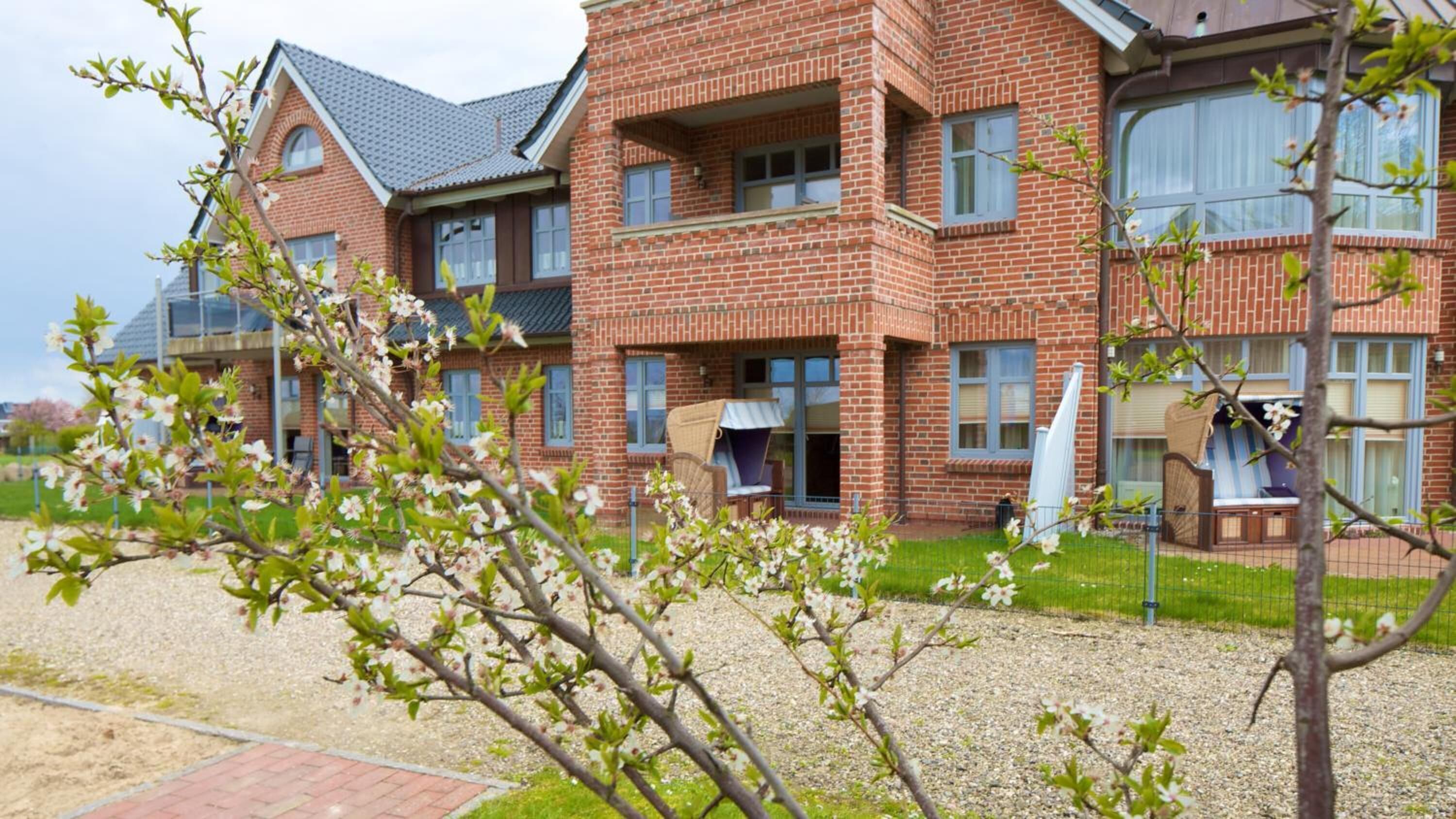 A multi-story brick building with balconies, a gravel driveway, and flowering trees.